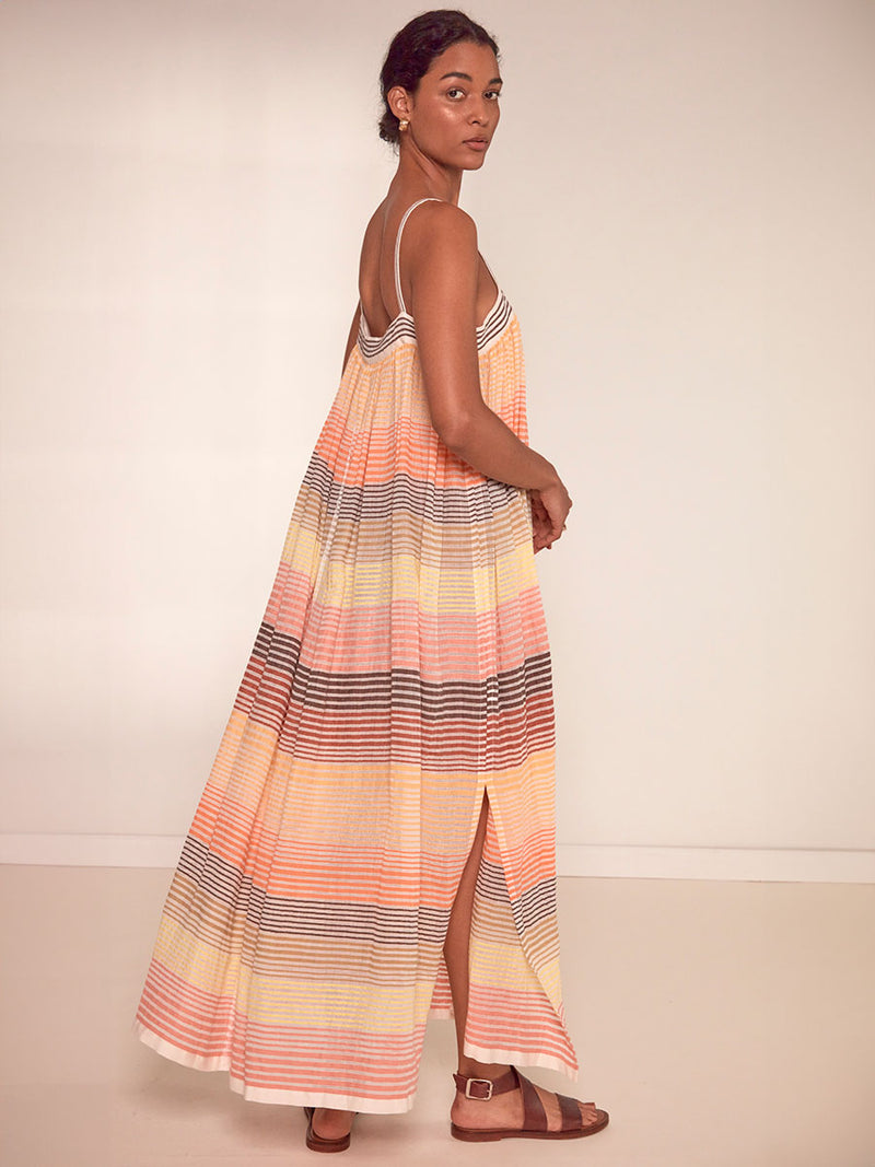 Side View of a Woman Standing Wearing lemlem Eda Flowy Dress Featuring continuous stripe pattern in warm yellow, orange and peach tones.