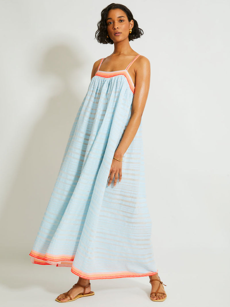 Woman Standing Wearing lemlem EDA Slip Dress featuring soft, variegated two-color stripe in sky blue and cream, complemented by placed bands of bright orange and tangerine.