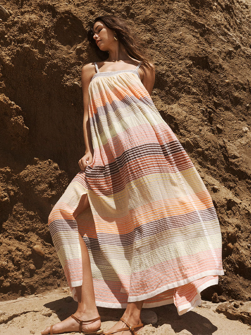 Woman Standing Wearing lemlem Eda Flowy Dress Featuring continuous stripe pattern in warm yellow, orange and peach tones.