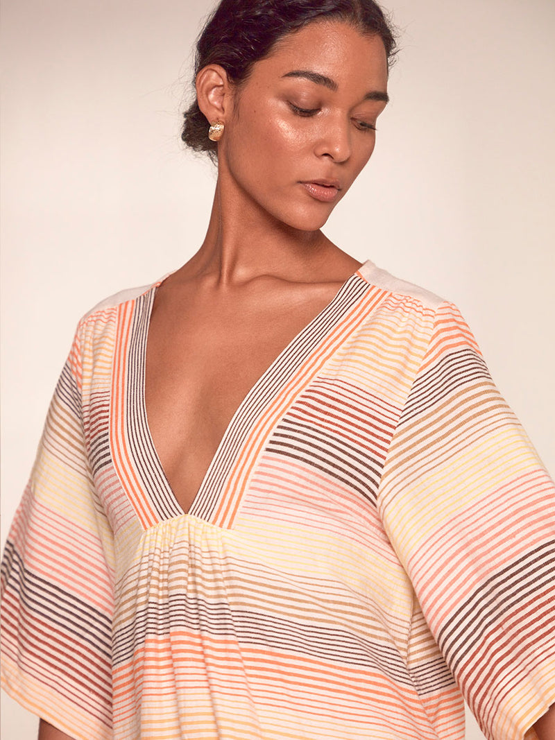 Close up on a Woman Standing Wearing lemlem Edna Dress Featuring continuous stripe pattern in warm yellow, orange and peach tones.