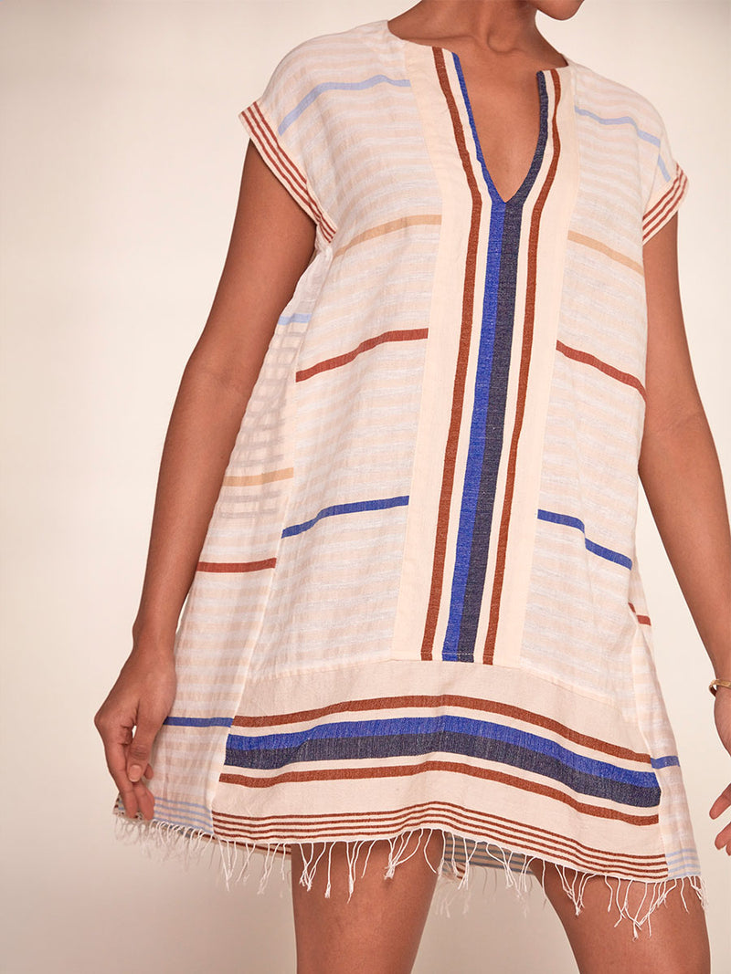 Close up on a Woman Standing Wearing lemlem Elina Caftan featuring striking bold stripe design in blue and brown hues on a neutral background