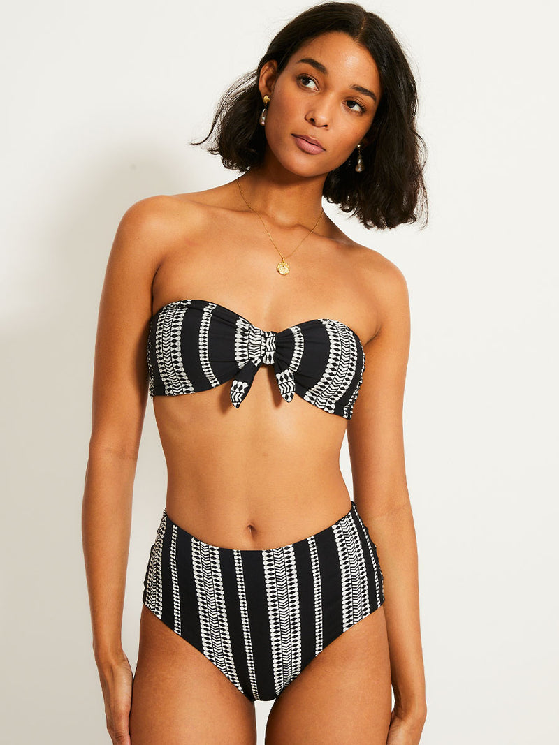 Woman standing wearing the Luchia bandeau bikini top and matching high waist bikini bottom in black with white graphic diamond and arrows.