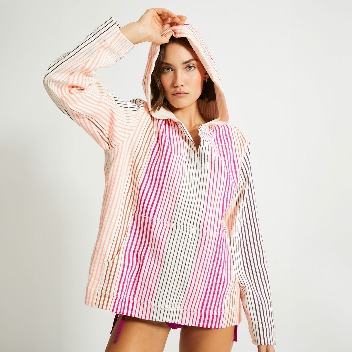 Woman standing wearing a lemlem hoodie in white with thin vertical pink, orange and brown stripes. 