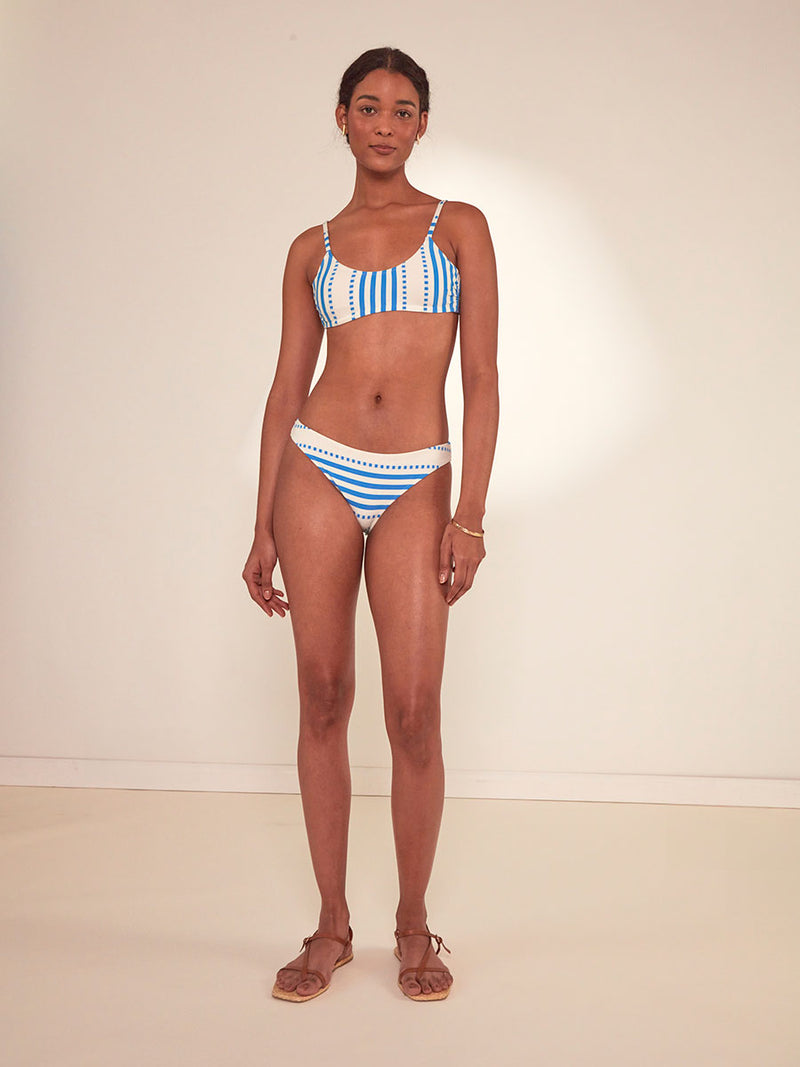 Woman Standing Wearing lemlem Meron Brief Bikini Bottom Featuring crisp white background and bright blue stripes and dots pattern and a matching Scoop Top