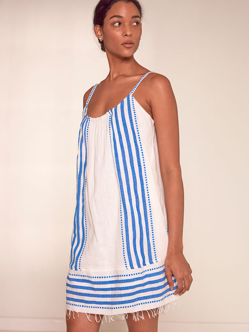 Side View of a Woman Standing Wearing lemlem Zina Swing Dress Featuring crisp white background and bright blue stripes and dots pattern