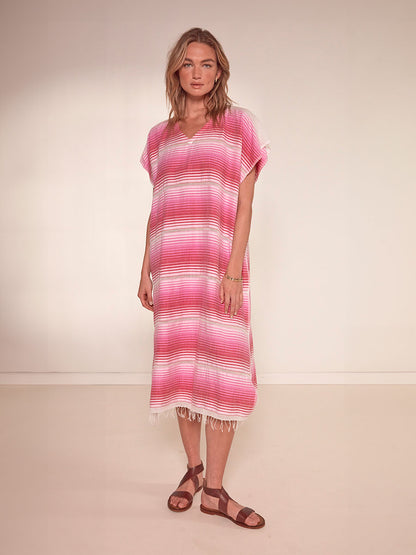 Woman Standing Wearing lemlem Dalila V Neck Caftan Dress featuring white, soft pink, and raspberry stripes that effortlessly blend into a stunning ombre effect
