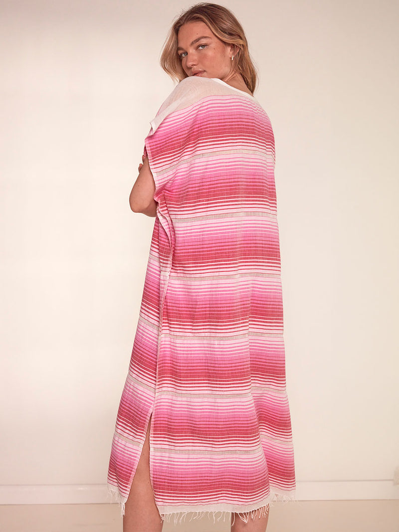 Back View of a Woman Standing Wearing lemlem Dalila V Neck Caftan Dress featuring white, soft pink, and raspberry stripes that effortlessly blend into a stunning ombre effect