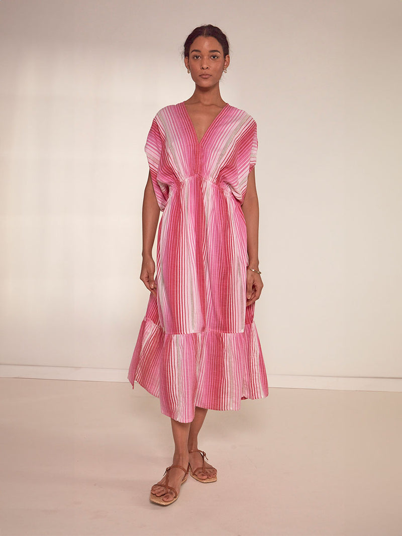 Woman Standing Wearing lemlem Leila Plunge Dress featuring white, soft pink, and raspberry stripes that effortlessly blend into a stunning ombre effect
