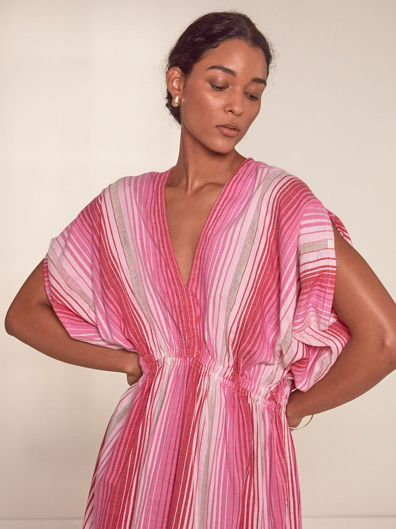 Woman Standing Wearing lemlem Leila Plunge Dress featuring white, soft pink, and raspberry stripes that effortlessly blend into a stunning ombre effect