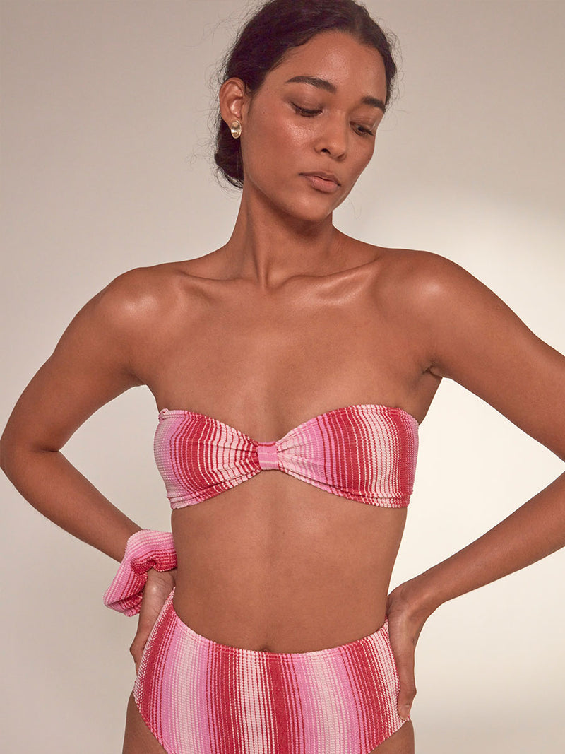 Close up on a Woman Standing Wearing Ennat Bandeau Top and lemlem Menen High Leg bikini bottom featuring striped fabric in ombre design in white, soft pink, and raspberry colors