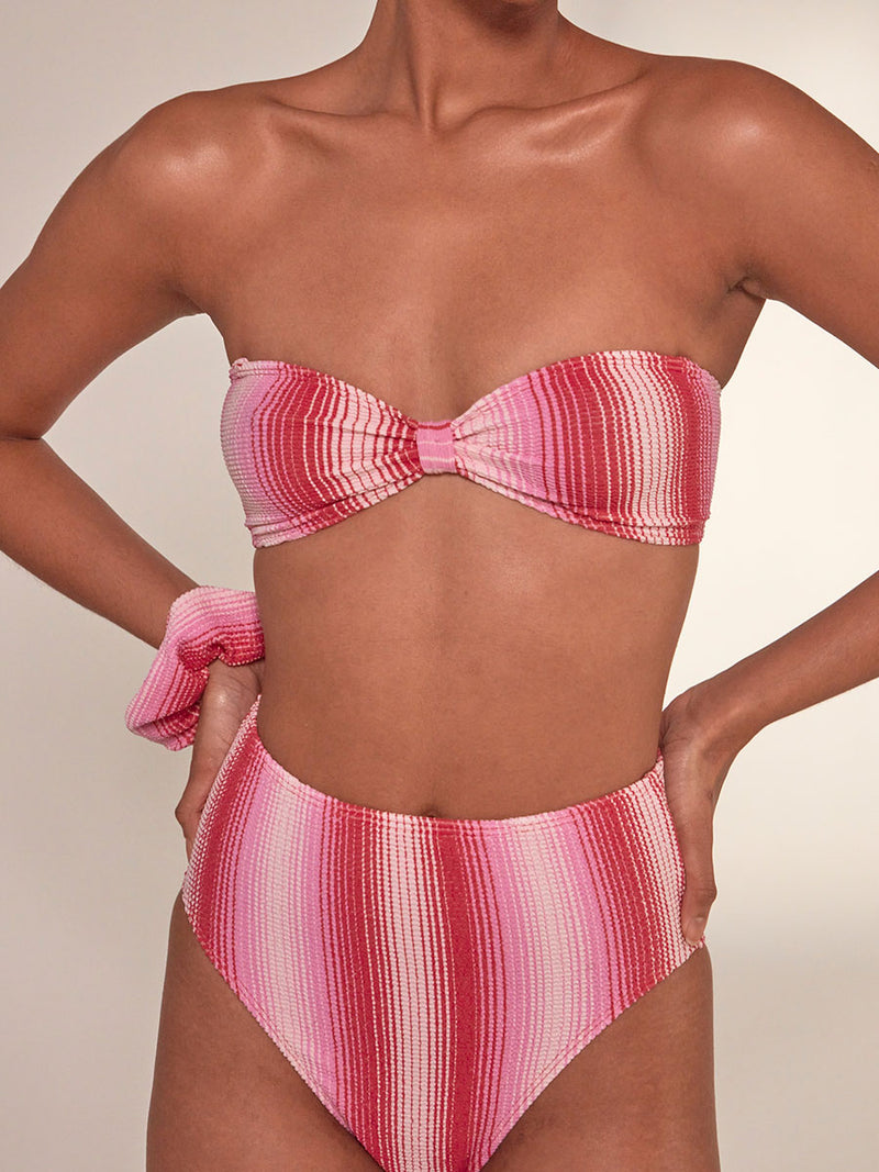 Woman Standing Wearing lemlem Lishan Scrunchie featuring striped fabric in ombre design in white, soft pink, and raspberry colors and matching bandeau top and high leg bikini bottom