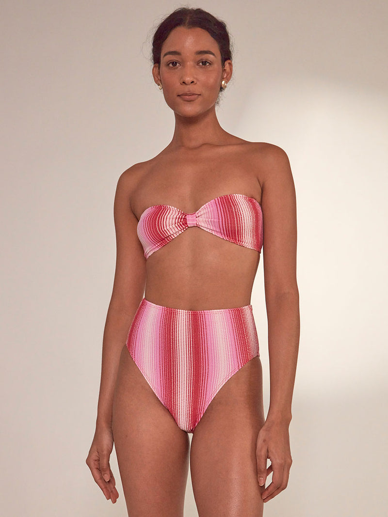 Woman Standing Wearing Ennat Bandeau Top and lemlem Menen High Leg bikini bottom featuring striped fabric in ombre design in white, soft pink, and raspberry colors