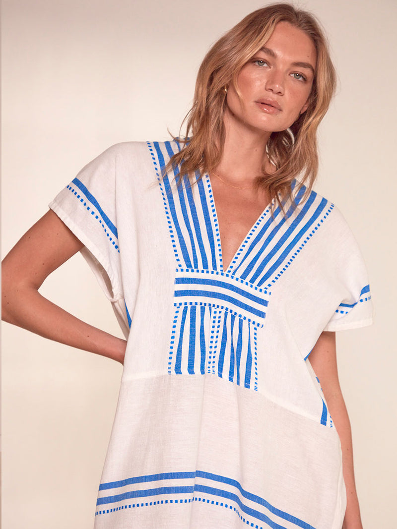 Close up on a Woman Standing Wearing lemlem Gasira V Neck Caftan Featuring crisp white background and bright blue stripes and dots pattern
