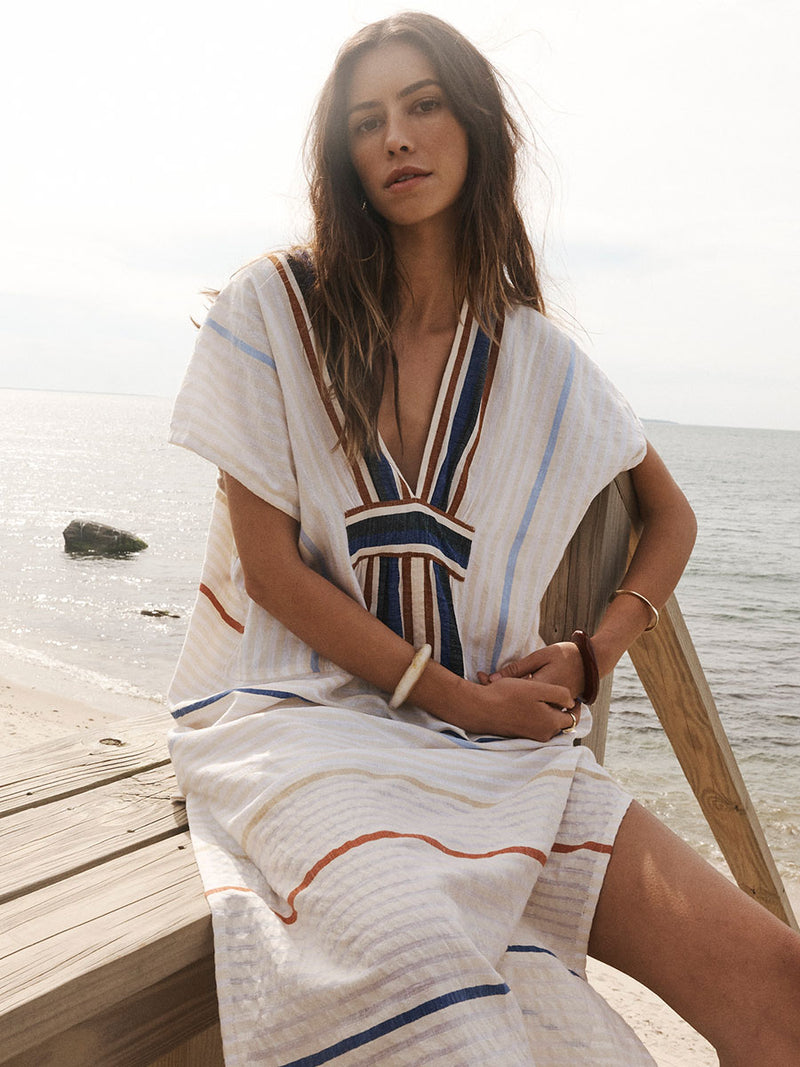 Woman Sitting on wooden stairs on the beach Wearing lemlem Gasira Caftan featuring striking bold stripe design in blue and brown hues on a neutral background