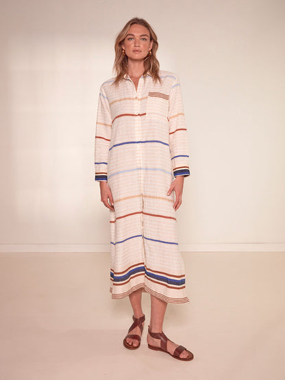 Woman Standing Wearing lemlem Anata Shirt Dress featuring striking bold stripe design in blue and brown hues on a neutral background