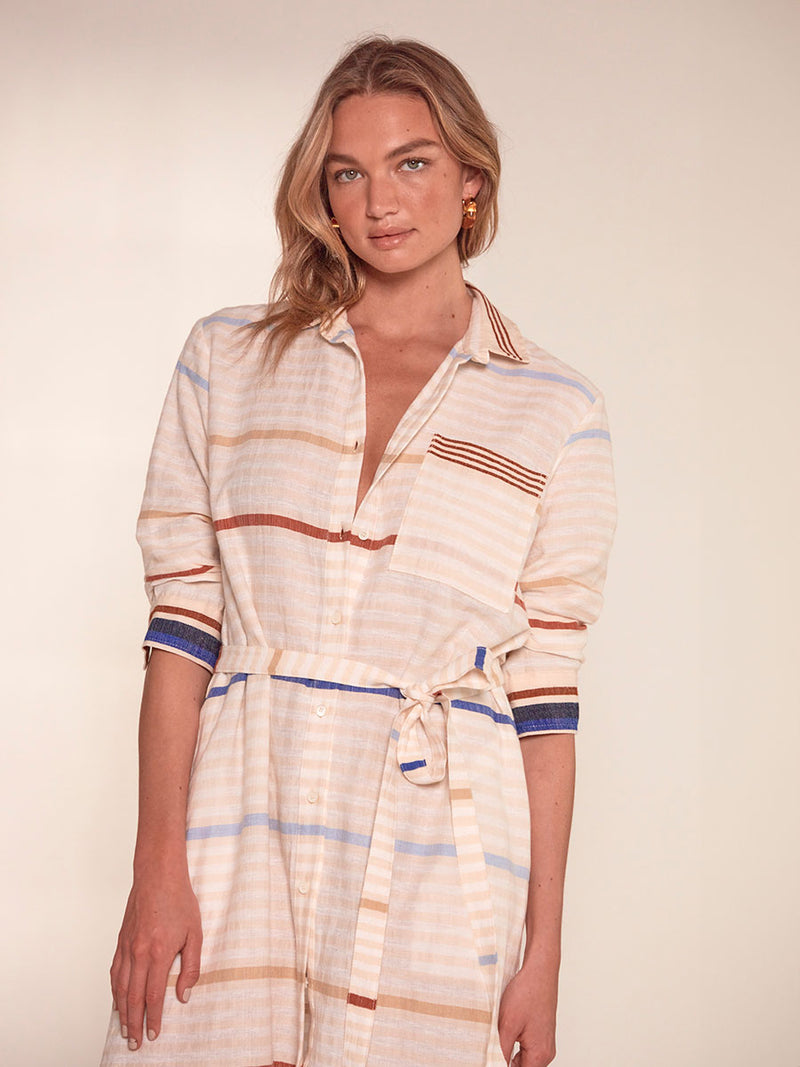  Close up on a Woman Standing Wearing lemlem Anata Shirt Dress featuring striking bold stripe design in blue and brown hues on a neutral background