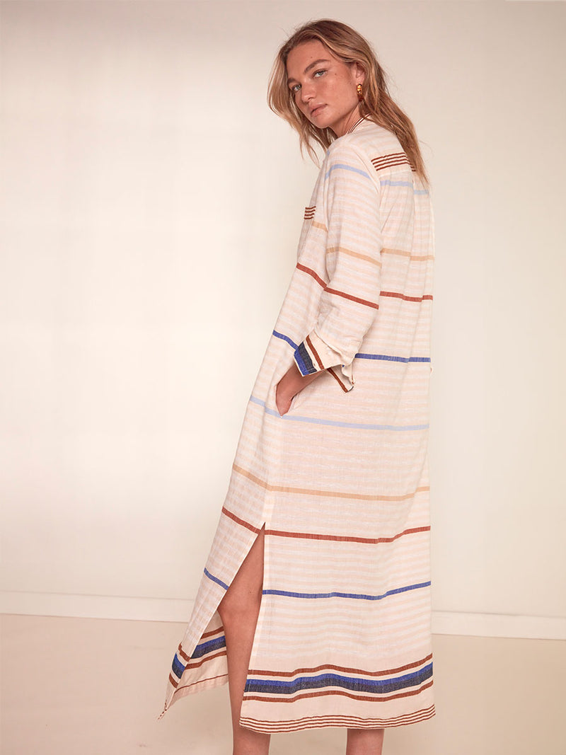 Side view of a Woman Standing Wearing lemlem Anata Shirt Dress featuring striking bold stripe design in blue and brown hues on a neutral background