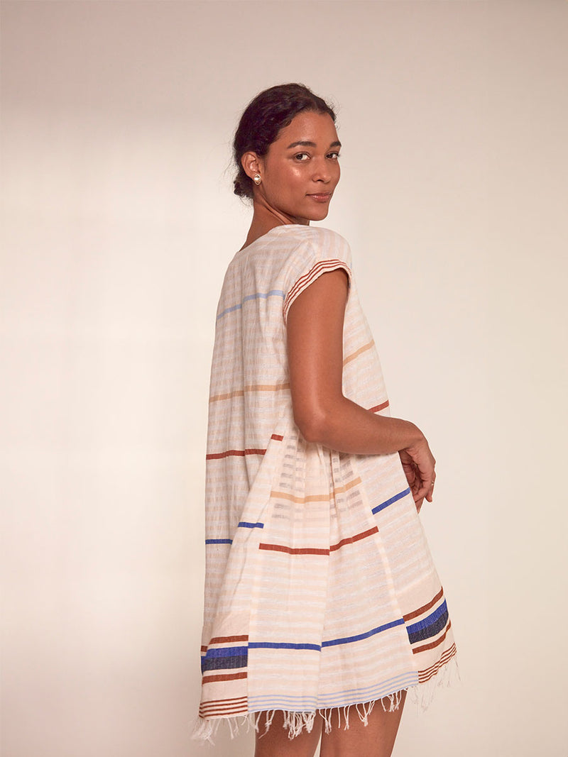 Side shot of Woman Standing Wearing lemlem Elina Caftan featuring striking bold stripe design in blue and brown hues on a neutral background