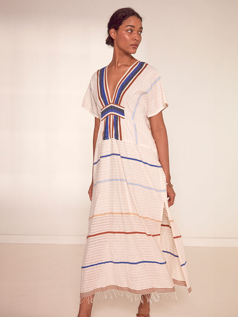 Woman Standing Wearing lemlem Gasira Caftan featuring striking bold stripe design in blue and brown hues on a neutral background
