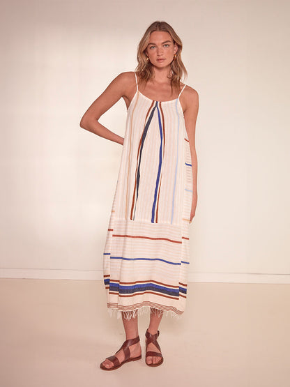 Woman Standing Wearing lemlem Nia Slip Dress featuring striking bold stripe design in blue and brown hues on a neutral background