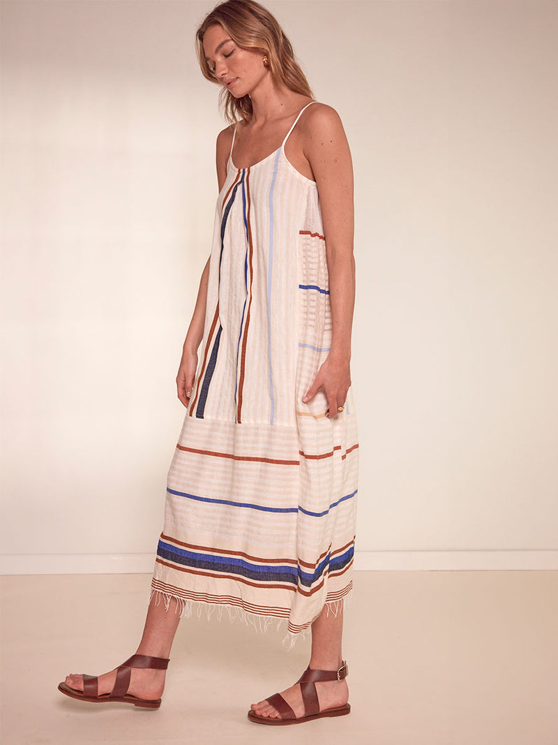 Side Shot of a Woman Standing Wearing lemlem Nia Slip Dress featuring striking bold stripe design in blue and brown hues on a neutral background