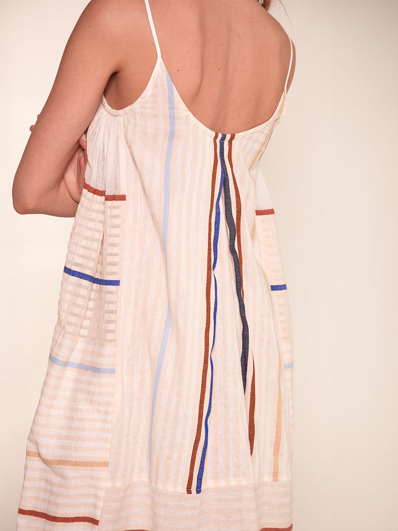 Back Close up on Woman's Back Standing Wearing lemlem Nia Slip Dress featuring striking bold stripe design in blue and brown hues on a neutral background