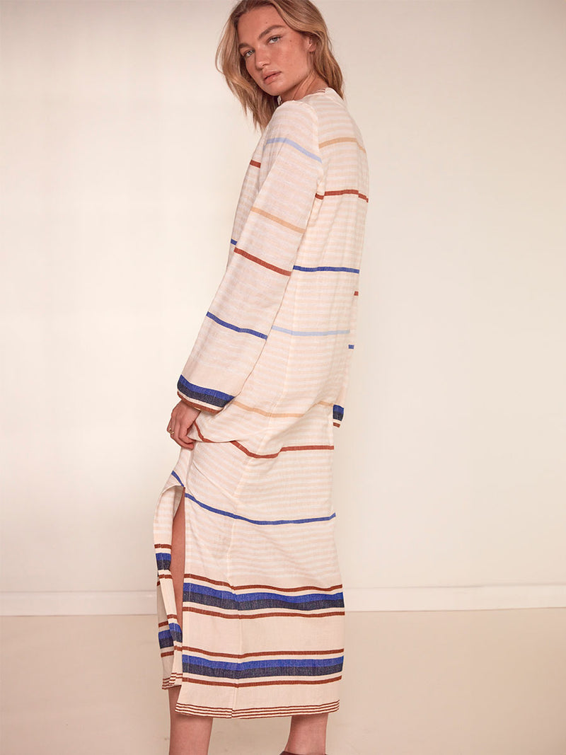 Side Shot of a Woman Standing Wearing lemlem Theodora Column Dress featuring striking bold stripe design in blue and brown hues on a neutral background