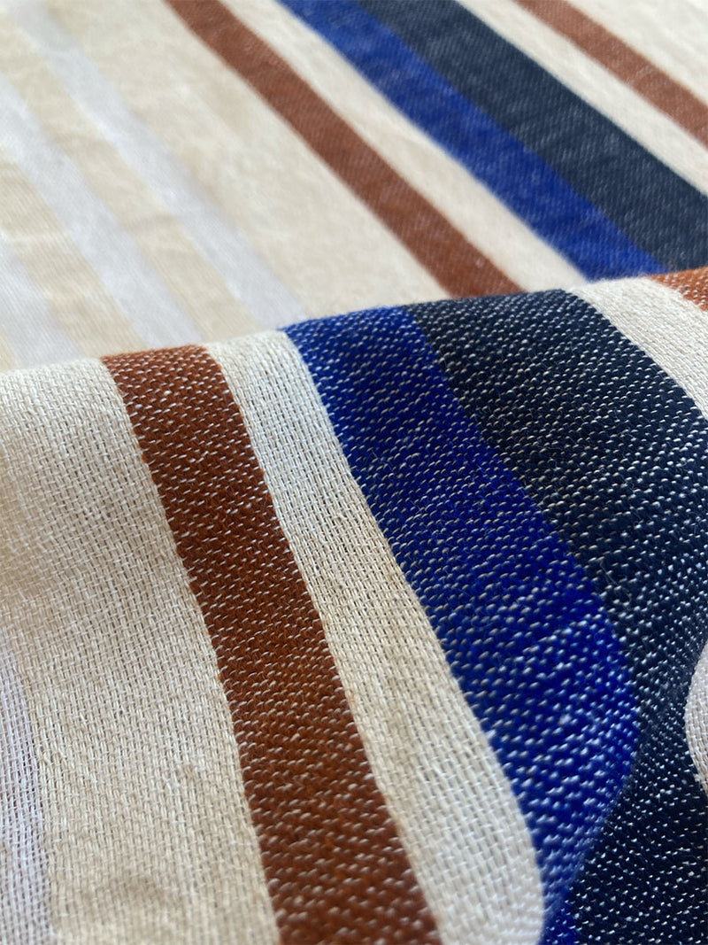 Close up on lemlem Hulet Vanilla Fabric featuring a neutral color palette and a striking bold stripe design in blue and brown hues on a neutral background