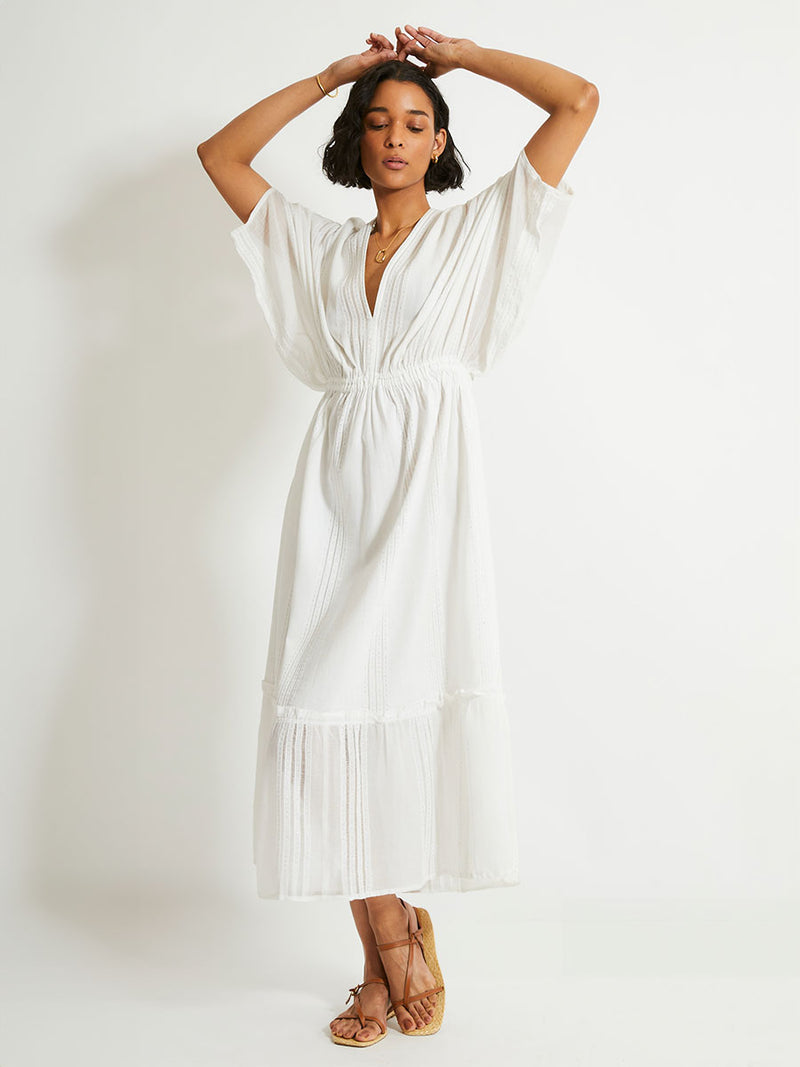 Woman standing wearing a white Abira plunge neck maxi dress with stitches of silver lurex