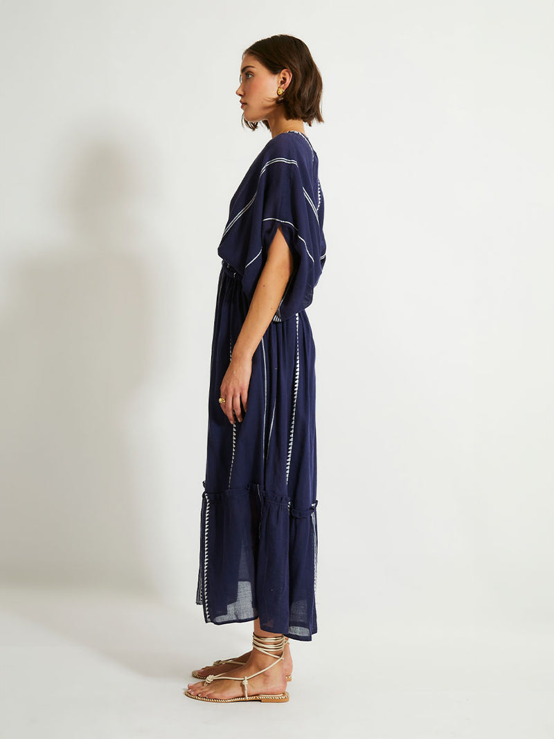 Side view of a woman standing with her hand on her hips wearing  the Nunu Plunge Neck dress in navy blue featuring a navy with white vertical stripes and graphic lines.