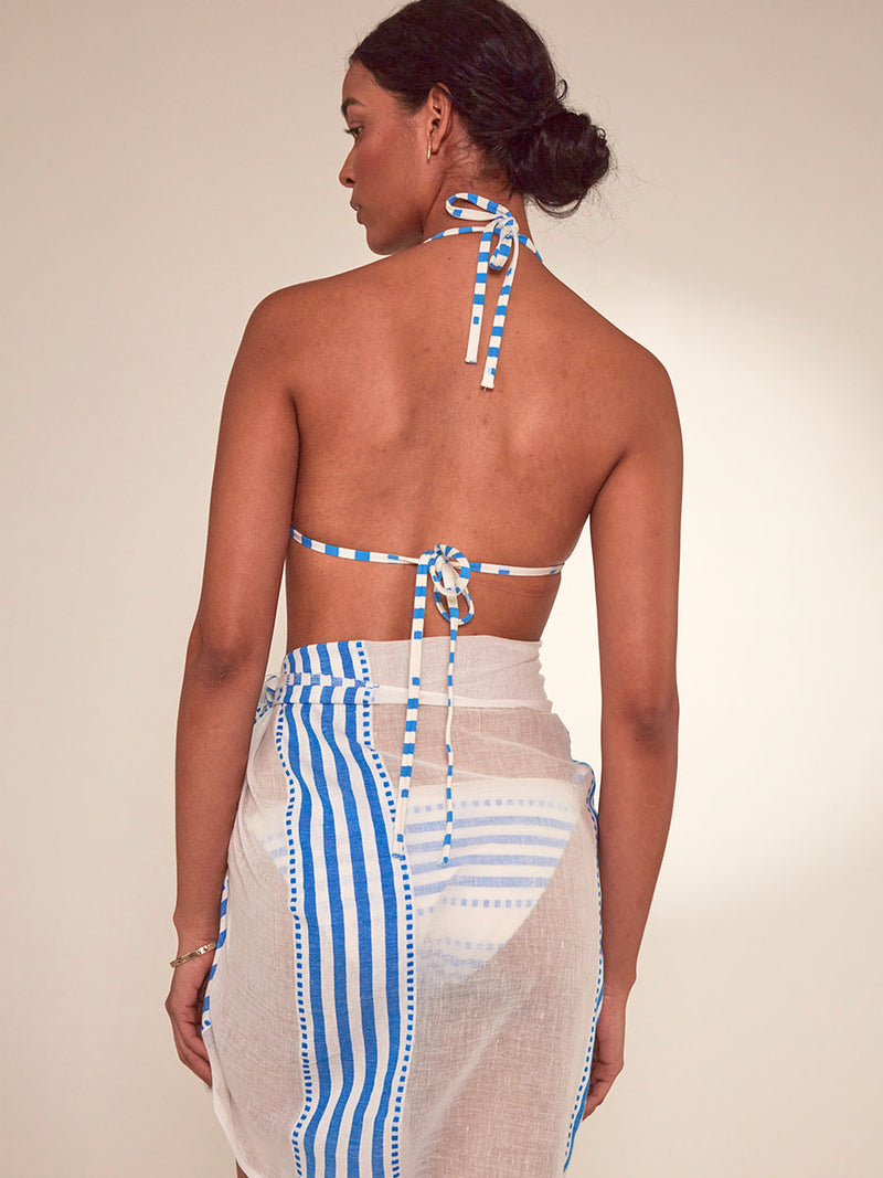 Back View of a Woman Standing Wearing lemlem Lema Sarong Featuring crisp white background and bright blue stripes and dots pattern, matching triangle top and string bikini bottom