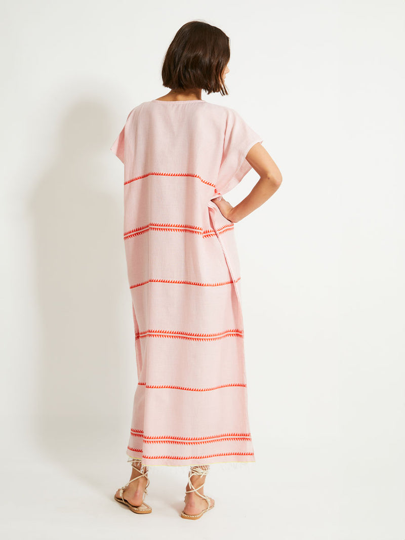 Back of a Woman Standing Wearing lemlem DALILA Caftan featuring vibrant orange red Tibeb pattern on a soft antique rose pink background, with a hint of citron tipping.