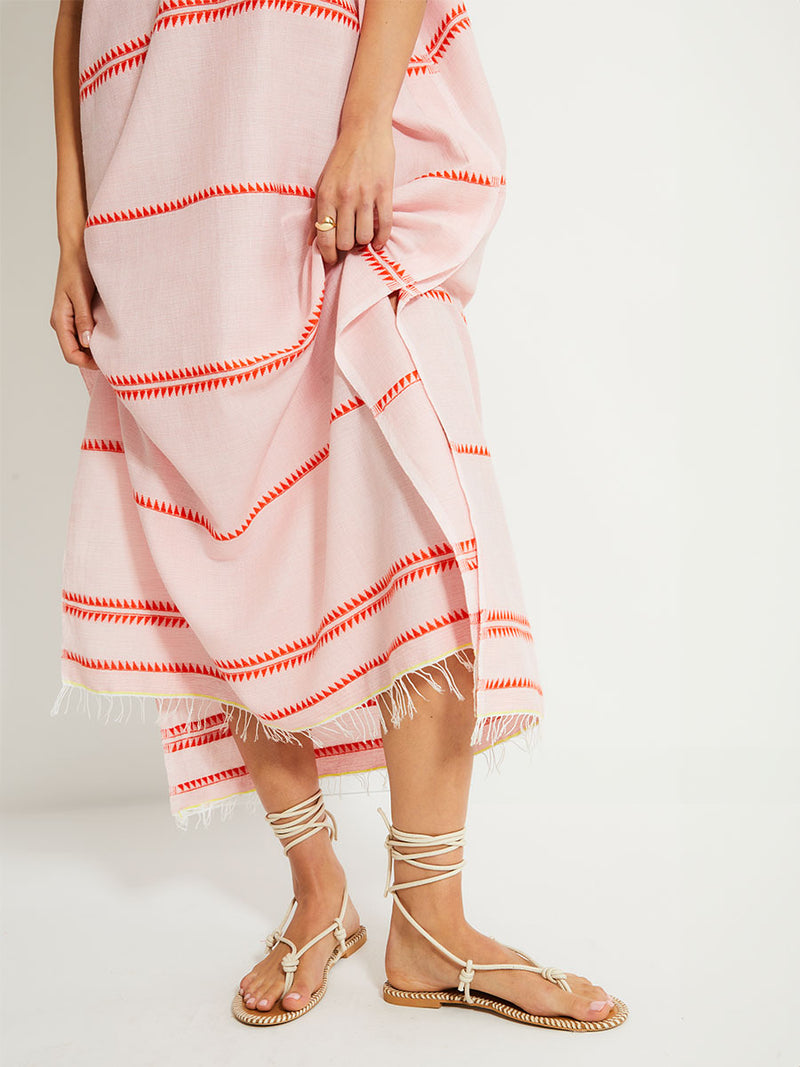 Close up on a Woman Standing Wearing lemlem DALILA Caftan featuring vibrant orange red Tibeb pattern on a soft antique rose pink background, with a hint of citron tipping.