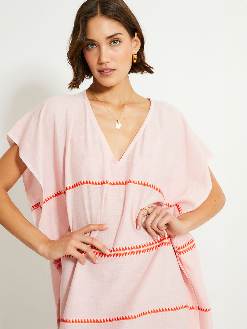 Close up on a Woman Standing Wearing lemlem DALILA Caftan featuring vibrant orange red Tibeb pattern on a soft antique rose pink background, with a hint of citron tipping.