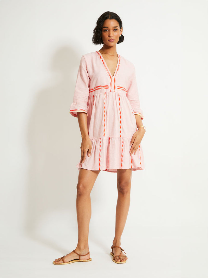 Woman Standing Wearing lemlem HANNA Dress featuring vibrant orange red Tibeb pattern on a soft antique rose pink background, with a hint of citron tipping.