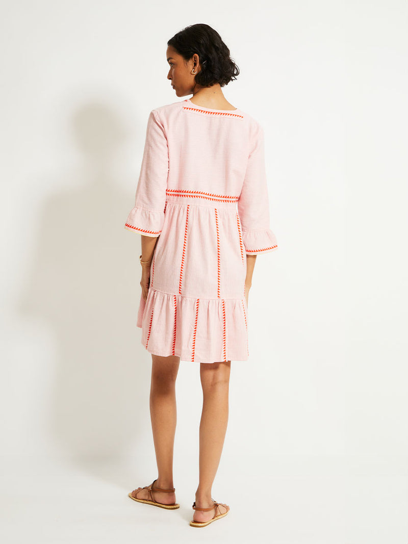 Back of a Woman Standing Wearing lemlem HANNA Dress featuring vibrant orange red Tibeb pattern on a soft antique rose pink background, with a hint of citron tipping.