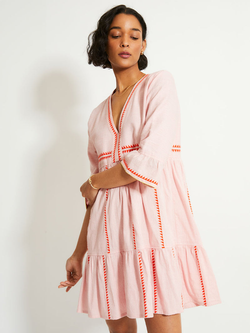 Woman Standing Wearing lemlem HANNA Dress featuring vibrant orange red Tibeb pattern on a soft antique rose pink background, with a hint of citron tipping.