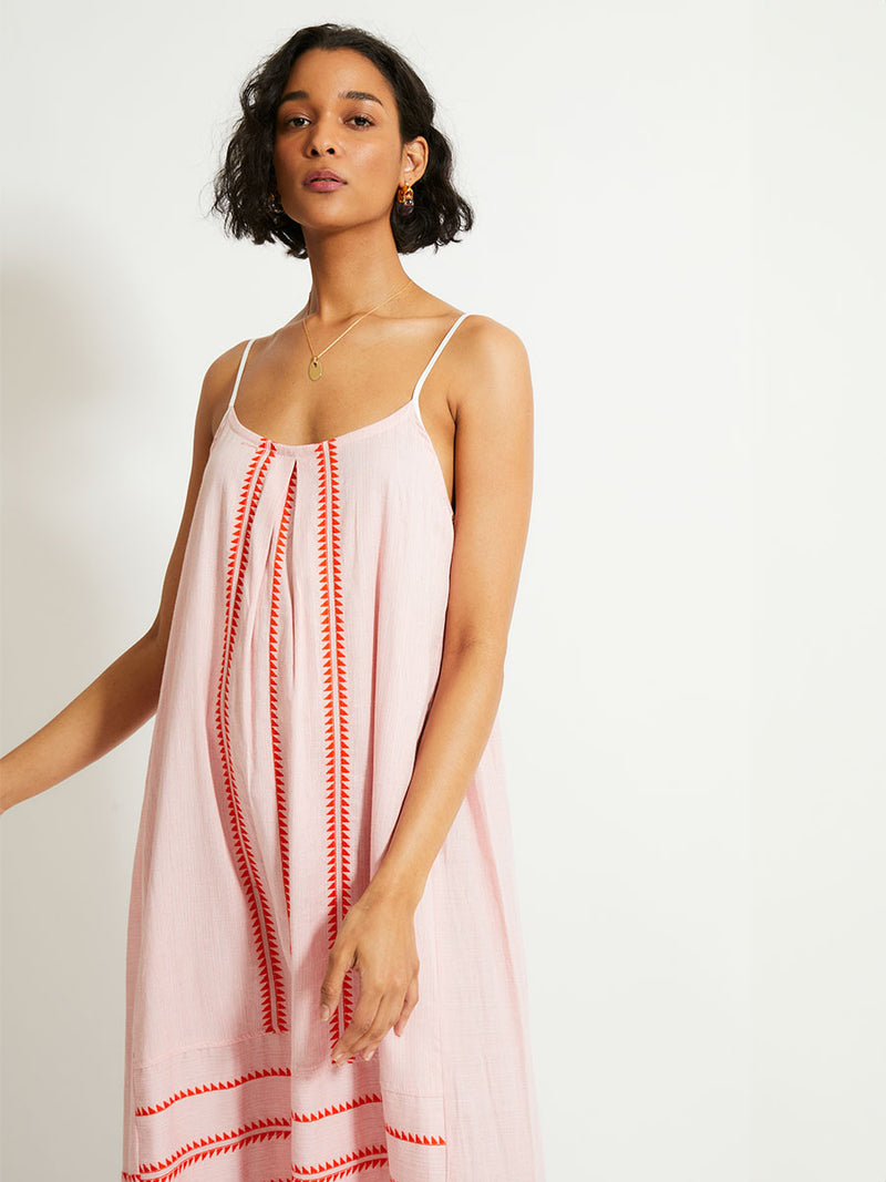 Close up on a Woman Standing Wearing  lemlem NIA Slip Dress featuring vibrant orange red Tibeb pattern on a soft antique rose pink background, with a hint of citron tipping.