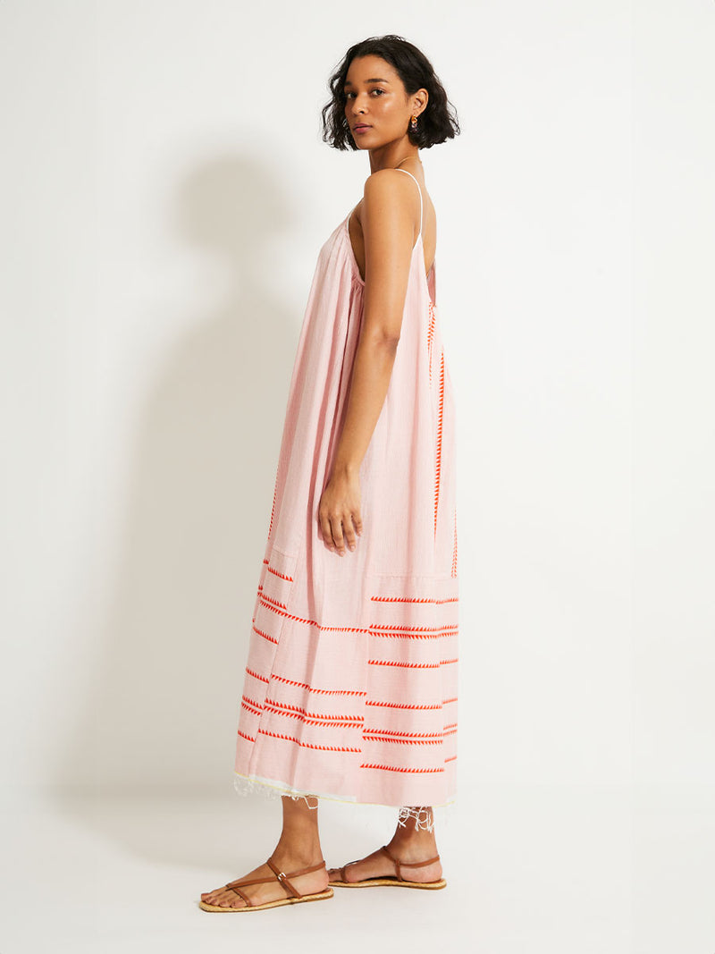 Side view of a Woman Standing Wearing  lemlem NIA Slip Dress featuring vibrant orange red Tibeb pattern on a soft antique rose pink background, with a hint of citron tipping.