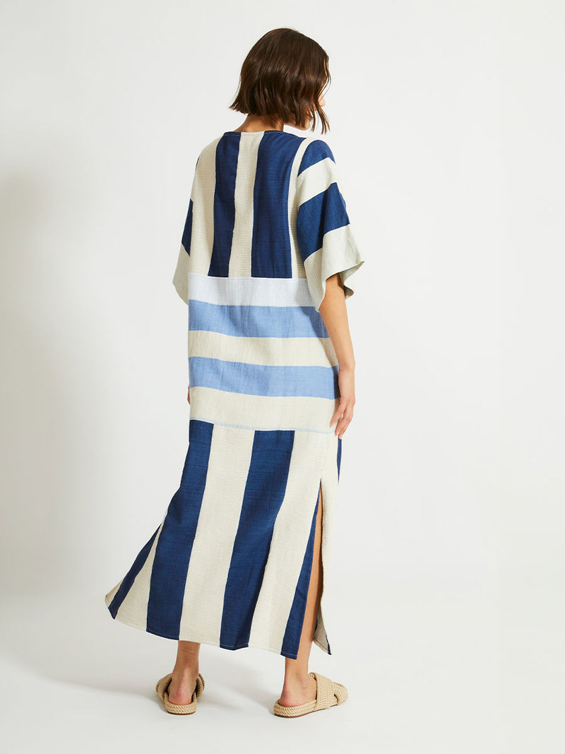 Back of a Woman Standing Wearing lemlem EDNA Dress featuring a striking color-block combination of three shades of blue, paired with bands of neutral cream and white background