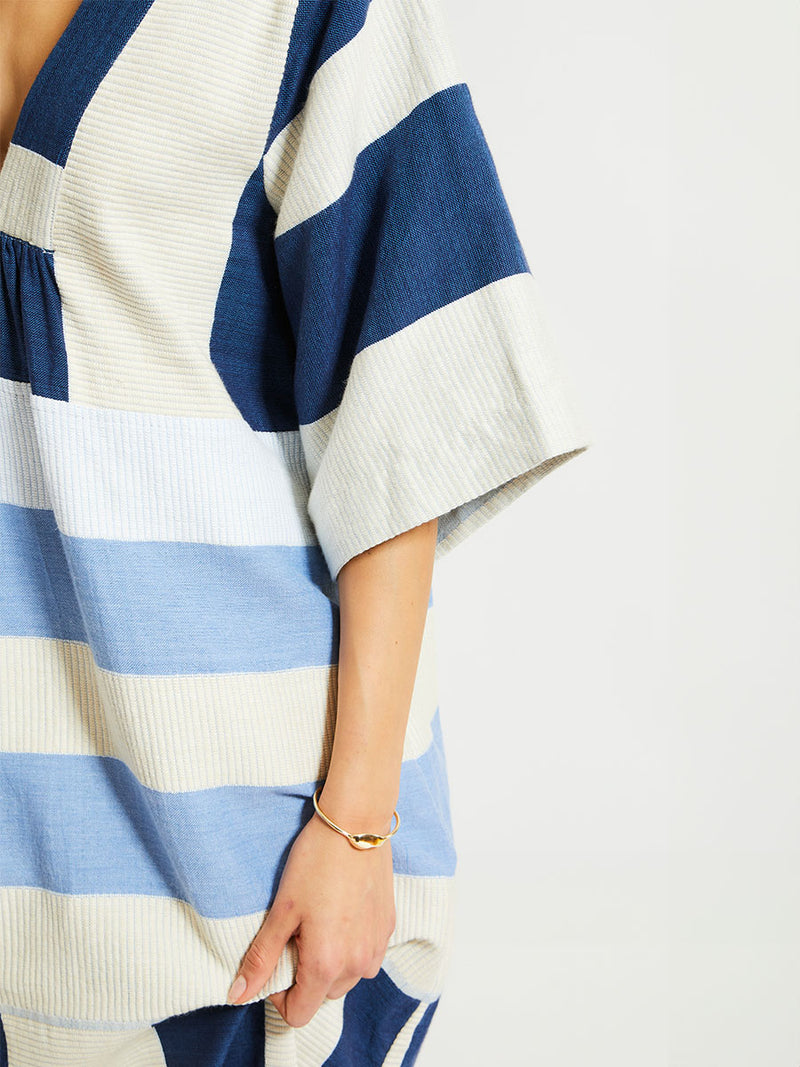 Close up on a Woman Standing Wearing lemlem EDNA Dress featuring a striking color-block combination of three shades of blue, paired with bands of neutral cream and white background