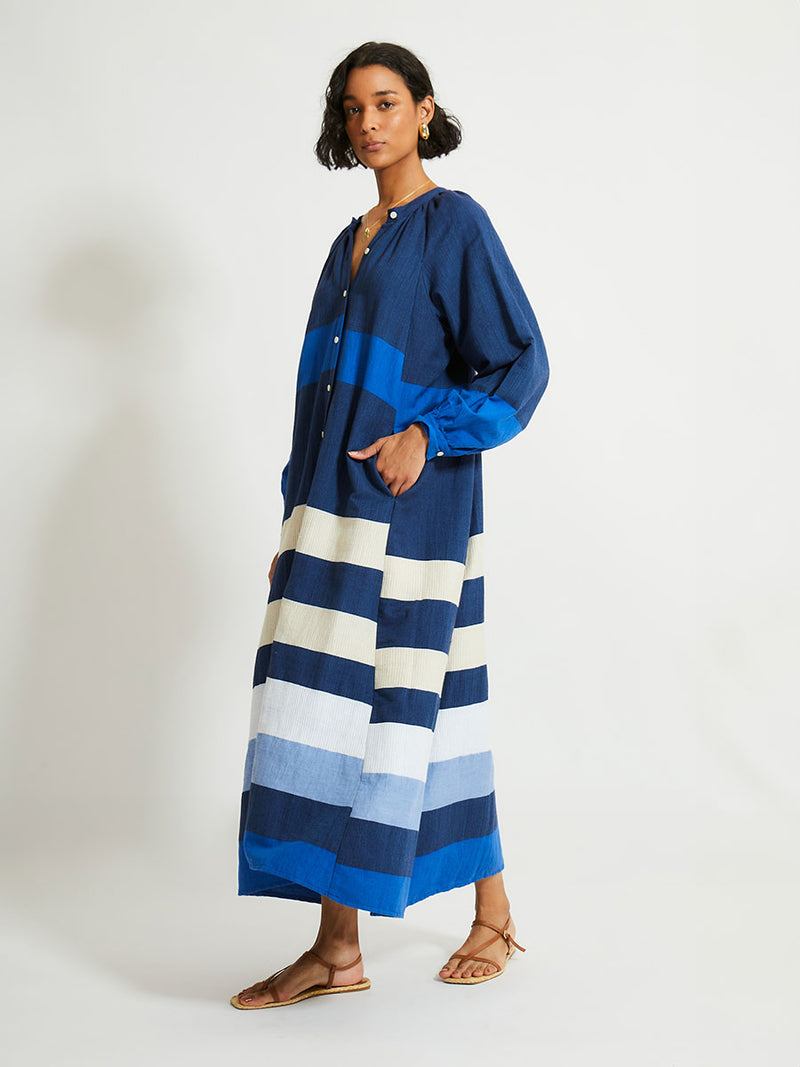 Side shot of a Woman Standing Wearing lemlem Makeda Dress featuring a striking color-block combination of three shades of blue, paired with bands of neutral cream and white