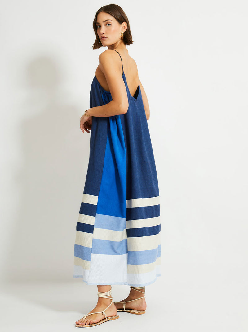 Side shot of a Woman Standing Wearing lemlem NIA Slip Dress featuring a striking color-block combination of three shades of blue, paired with bands of neutral cream and white