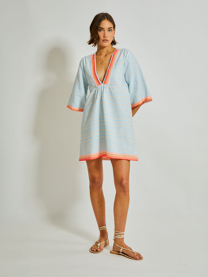 Woman Standing Wearing lemlem Belkis Caftan featuring soft, variegated two-color stripe in sky blue and cream, complemented by placed bands of bright orange and tangerine.