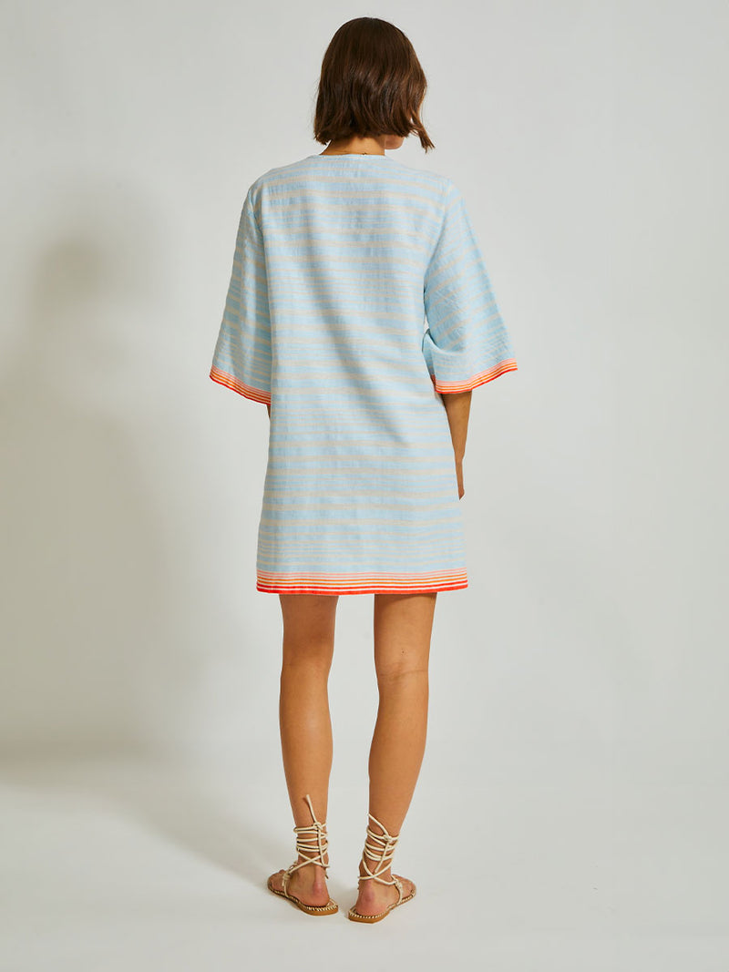 Back of a Woman Standing Wearing lemlem Belkis Caftan featuring soft, variegated two-color stripe in sky blue and cream, complemented by placed bands of bright orange and tangerine.