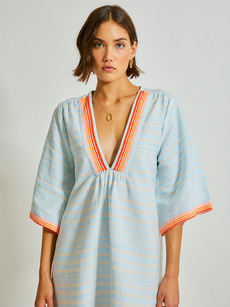 Woman Standing Wearing lemlem Belkis Caftan featuring soft, variegated two-color stripe in sky blue and cream, complemented by placed bands of bright orange and tangerine.