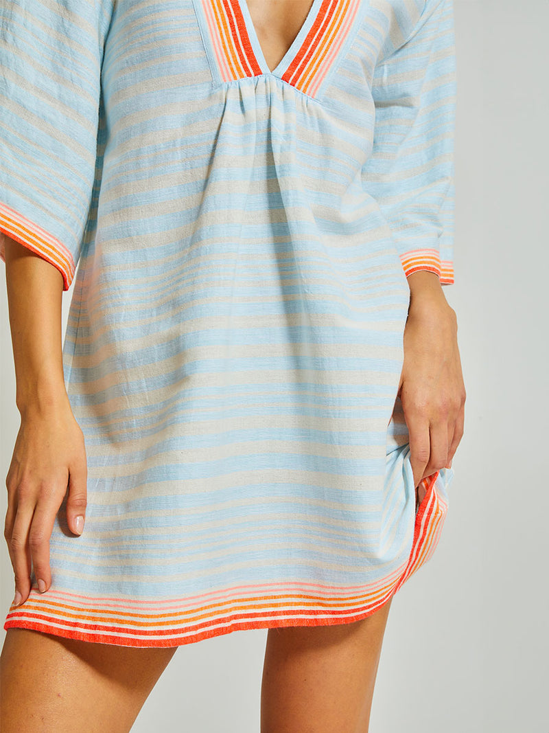 close up on a Woman Standing Wearing lemlem Belkis Caftan featuring soft, variegated two-color stripe in sky blue and cream, complemented by placed bands of bright orange and tangerine.