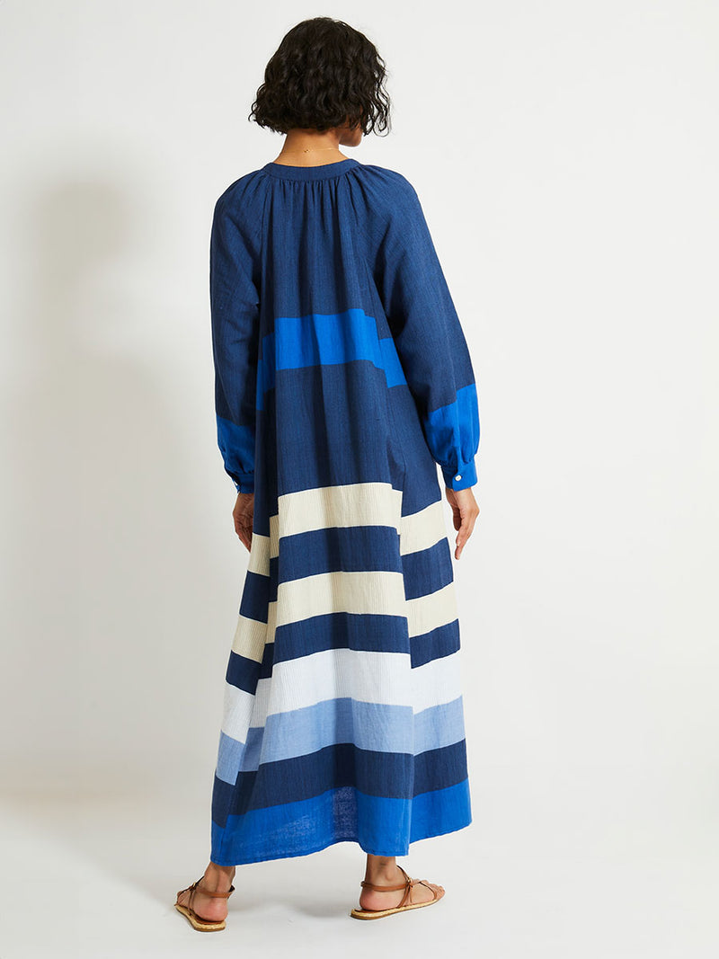 Back of a Woman Standing Wearing lemlem Makeda Dress featuring a striking color-block combination of three shades of blue, paired with bands of neutral cream and white