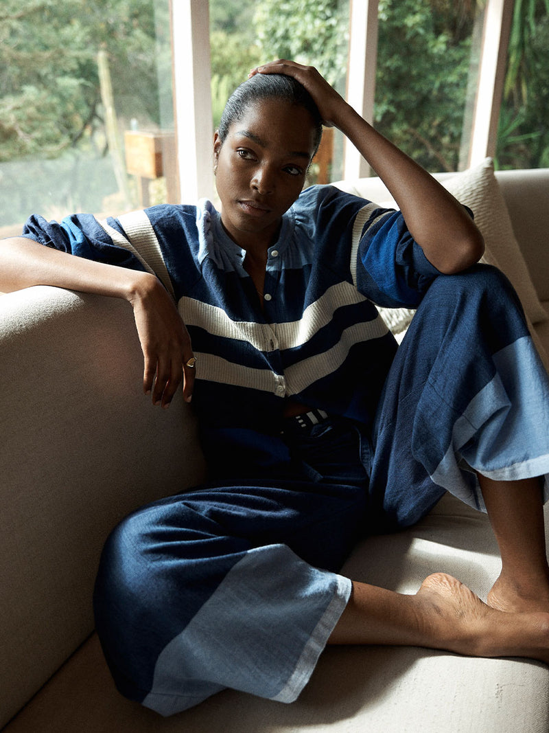 Woman Sitting on the couch Wearing lemlem MITA Blouse featuring a striking color-block combination of three shades of blue, paired with bands of neutral cream and white and matching pants