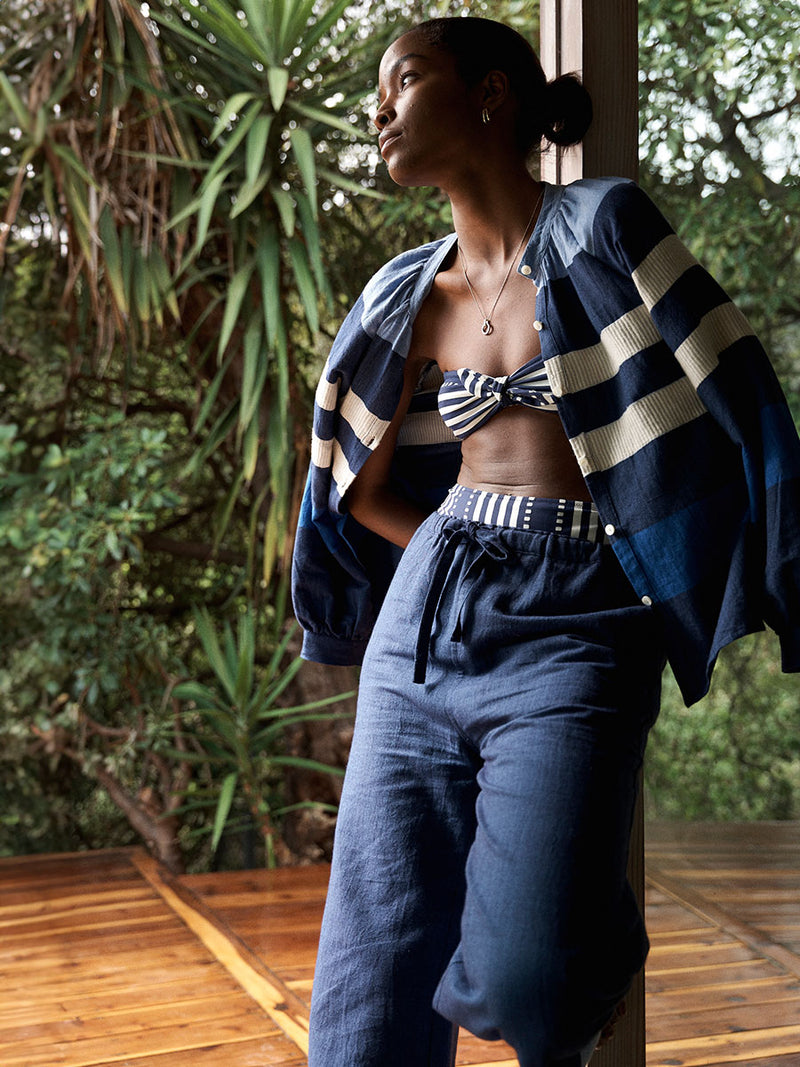 Woman Standing in the tree house Wearing lemlem MITA Blouse featuring a striking color-block combination of three shades of blue, paired with bands of neutral cream and white and matching pants